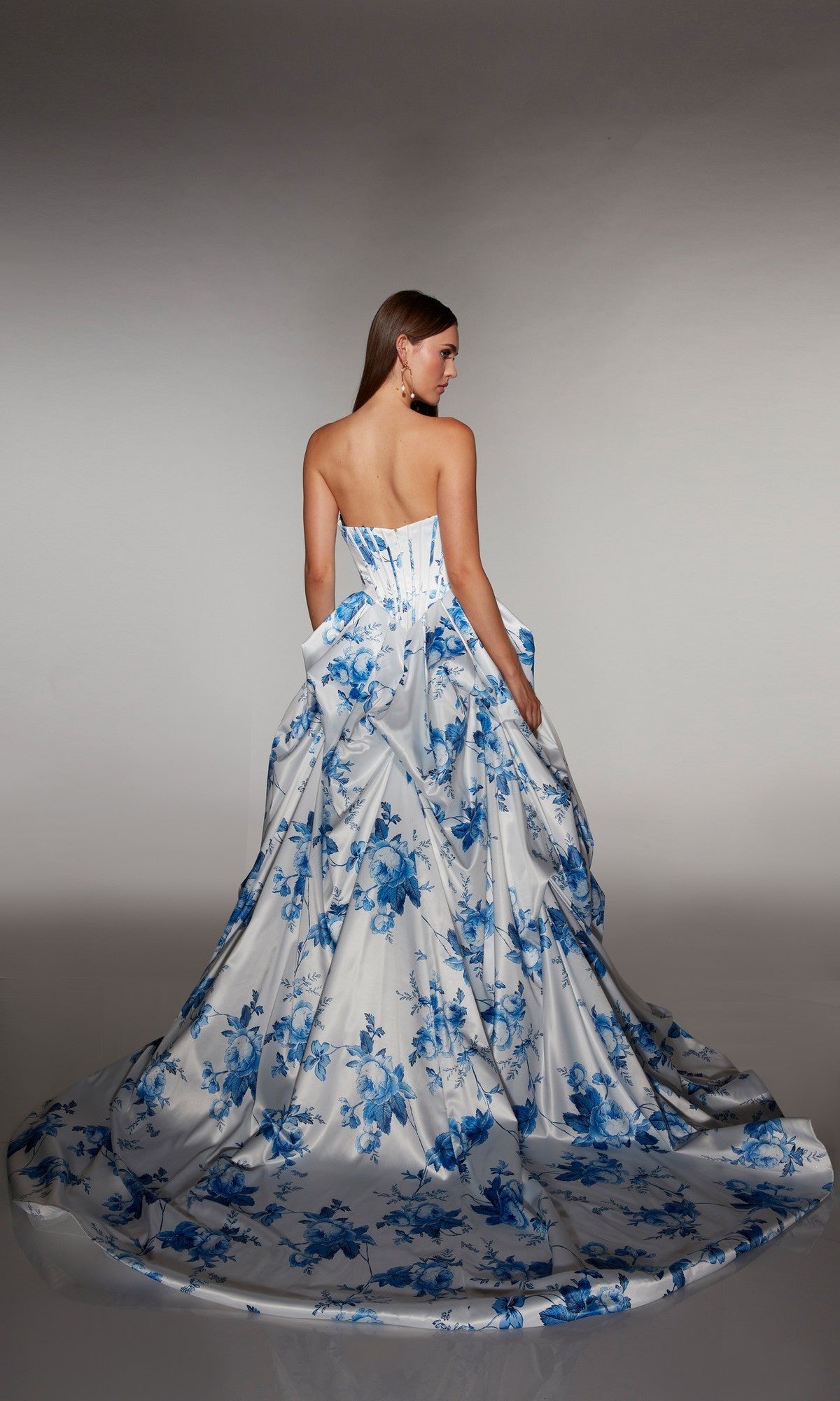 A woman stands with her back to the camera, wearing the Alyce 62213 long prom dress. This strapless gown features a corset bodice and a flowing white skirt decorated with elegant blue floral patterns.